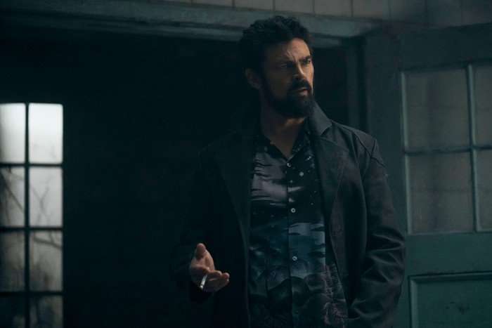 Karl Urban as Billy Butcher in The Boys, wearing a black and white shirt and a black coat, and holding a cigarette, stood in a doorway.