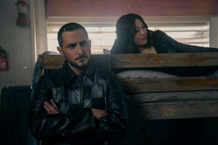 Tomer Capone as Frenchie and Karen Fukuhara as Kimiko in The Boys. She is lying on a bunkbed and he is stood next to it with his arms folded.
