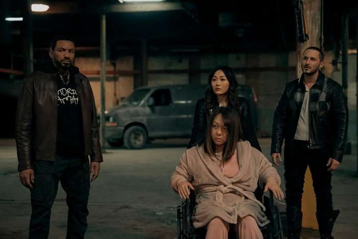 Laz Alonso as Mother's Milk, Karen Fukuhara as Kimiko, Tomer Capone as Frenchie and Naoko Mori as Golden Geisha in The Boys. MM, Kimiko and Frenchie are stood in a warehouse with a van parked behind them, while Holden Geisha is wearing a dressing gown and sat in a chair in front of them.