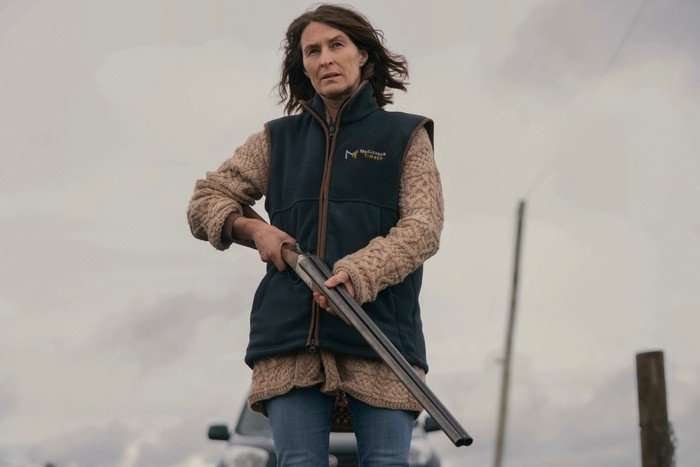 THE_DARK_HelenBaxendale Helen Baxendale as Bethany Morgan standing outside on a grey day and holding a rifle in her hands, looking stressed as she also wears a gilet and shirt.