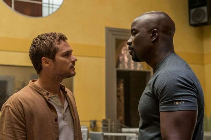 Finn Jones and Mike Colter as Danny Rand and Luke Cage. They are stood opposite each other in a domestic setting, having a conversation
