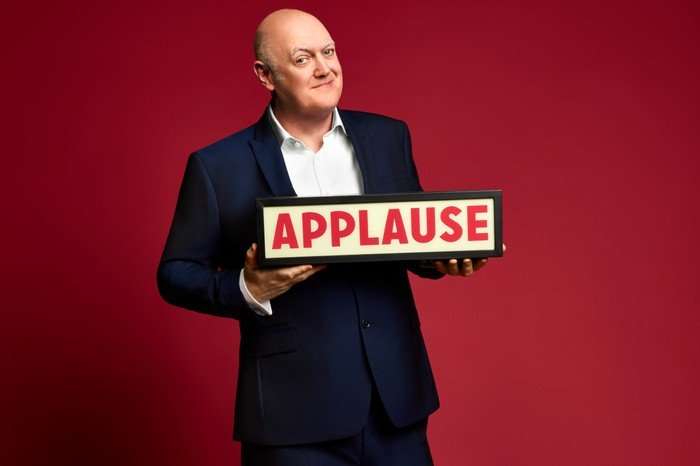 Dara &Oacute; Briain in a navy suit smiles while holding an illuminated &ldquo;APPLAUSE&rdquo; sign against a solid red studio background.