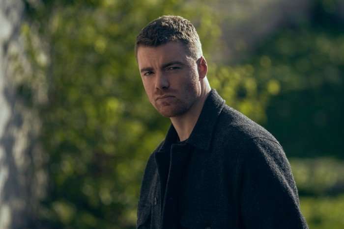 Gabriel Basso as Peter Sutherland in The Night Agent, wearing black and stood in front of a tree.