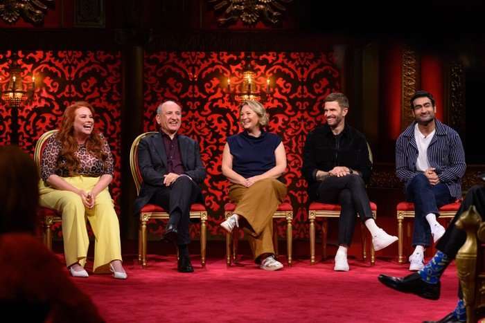 Amy Gledhill, Armando Iannucci, Joanna Page, Joel Dommett and Kumail Nanjiani sitting on chairs in the Taskmaster studio, all smiling and laughing.