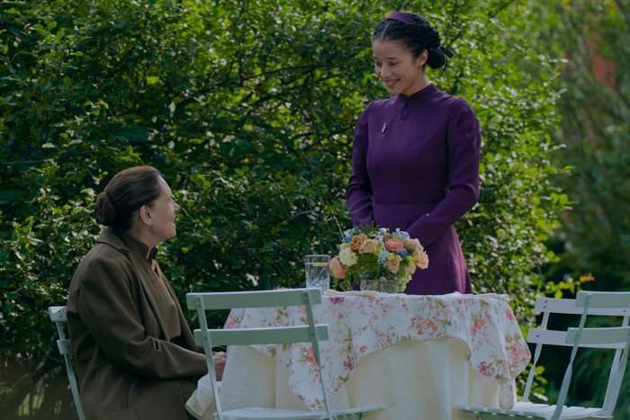 Ann Dowd sitting at a table in a garden and Chase Infiniti approaching with a smile on her face in The Testaments.