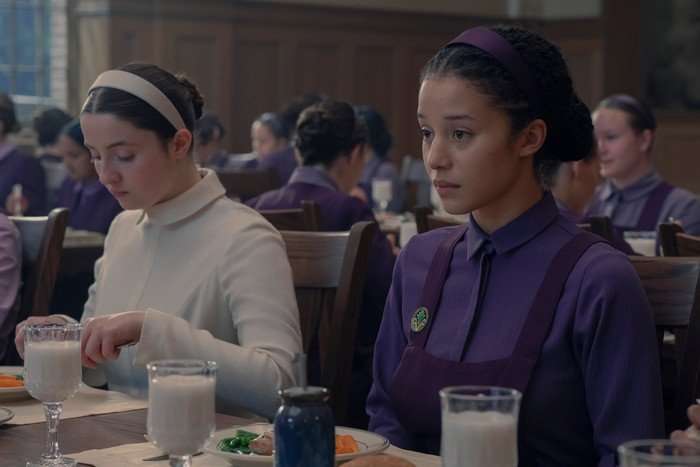 Lucy Halliday and Chase Infiniti in The Testaments sitting at their lunch table, eating and looking nervously.