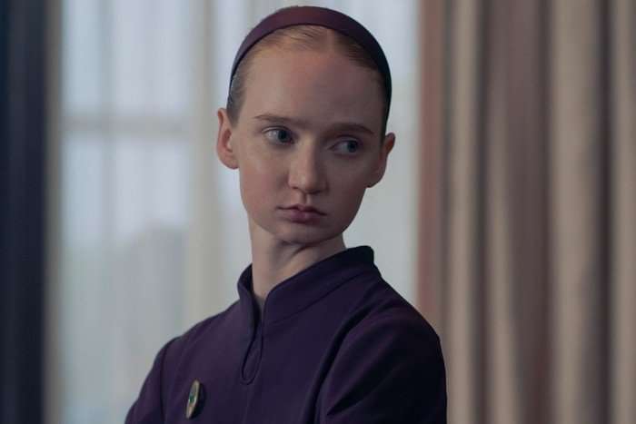 Isolde Ardies in The Testaments standing in the middle of a room and turning around, looking serious, dressed in a purple dress and headband.
