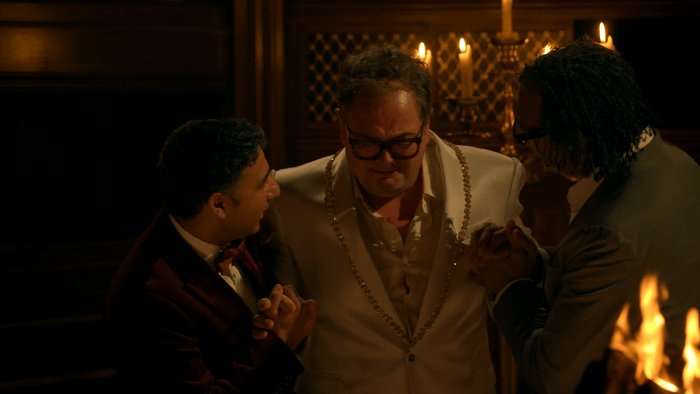 The Celebrity Traitors' Alan Carr, David Olusoga and Nick Mohammed huddle by candlelight in a dark, wood-panelled room, clasping hands with tense, conspiratorial expressions.