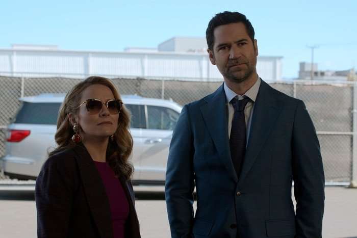 Becki Newton as Lorna Crane and Manuel Garcia-Rulfo as Mickey Haller in The Lincoln Lawyer, stood outside together with a car behind them. She is wearing a red top, black jacket and sunglasses, and he is wearing a blue suit.