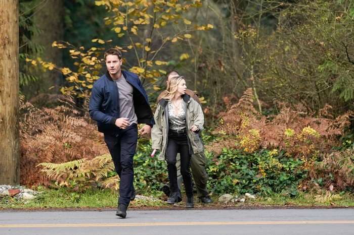 Justin Hartley as Colter Shaw and Sydney Scotia as Angie in Tracker season 2 running across a main road that's surrounded by the woods.