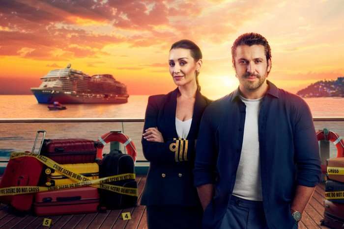 A man stands with his hands in his pockets a woman to his left and slightly behind him stands with his arms crossed. They both smile ahead with a sunset behind them.