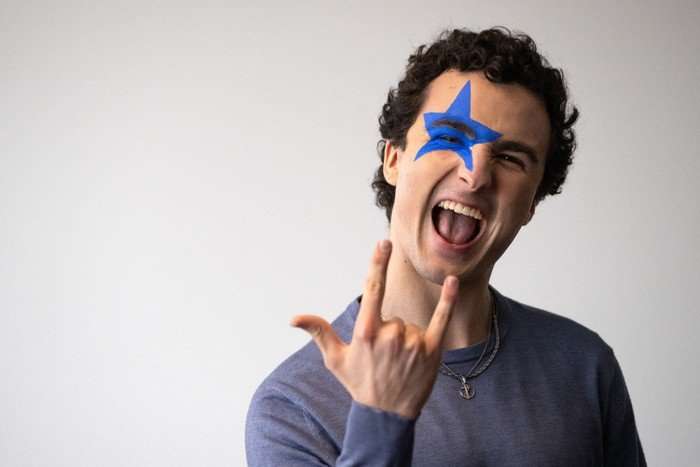 Eurovision-2026-austria COSMÓ looking ahead with his mouth ajar, a blue star painted across one eye and making the rock and roll symbol with one hand.