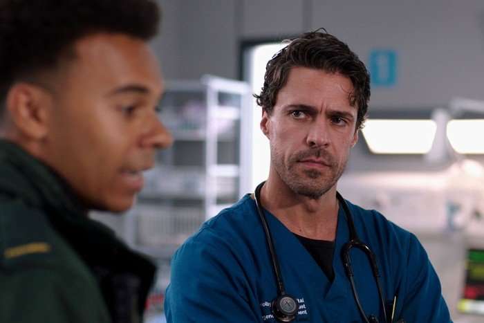 A man dressed in scrubs and a stethoscope around his neck looks seriously at another man dressed as a paramedic and out of focus.