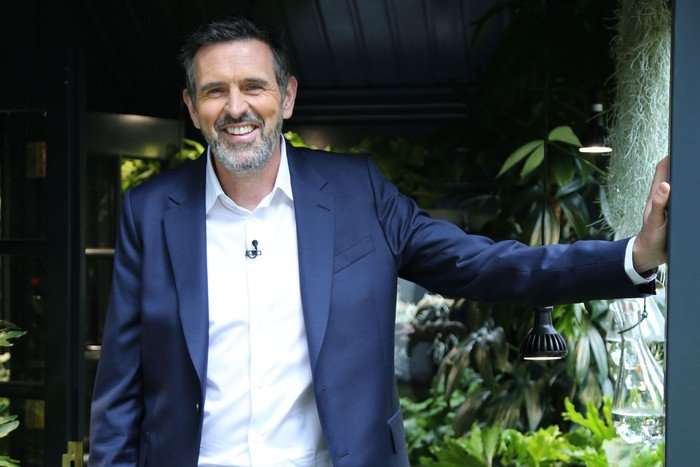 Adam Frost wearing a white shirt and blue blazer, stood in a doorway smiling.
