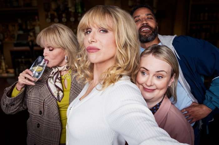 A group of four adults pose closely together in what appears to be a bar setting. In the foreground, a blonde woman with shoulder-length wavy hair and bangs faces the camera with a confident expression, wearing a light-colored top. Behind her to the right, a smiling man leans in casually. In front of him, a woman with light hair pulled back rests her head against the central figure&rsquo;s shoulder, smiling warmly. On the left, an older woman in a patterned jacket and scarf drinks from a glass with a lemon slice, looking slightly away from the camera. The background is softly blurred, showing shelves of bottles and a cozy bar atmosphere.