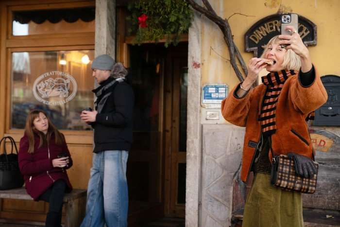 THE FOUR SEASONS, SEASON 2 A blonde woman stands outside a restaurant in a picturesque European street, taking a selfie while eating a slice of pizza. She is wearing a rust-coloured jacket, a striped scarf and a crossbody bag, smiling brightly at her phone. Other people stand and sit nearby with takeaway coffees, while festive greenery and rustic signage decorate the building behind her.