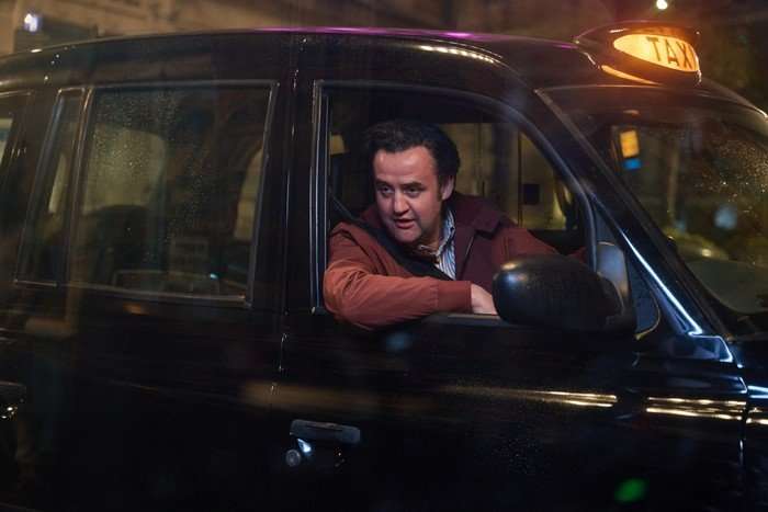 Daniel Mays as John Worboys in Believe Me driving a black cab and leaning out of his open window to speak to someone on the pavement.