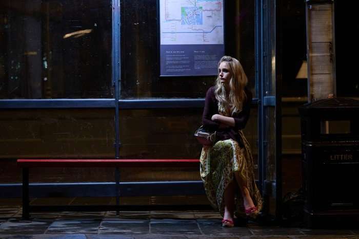 Miriam Petche as Carrie Symonds in Believe Me sitting at a bus stop, crossing her legs as she waits in the night.