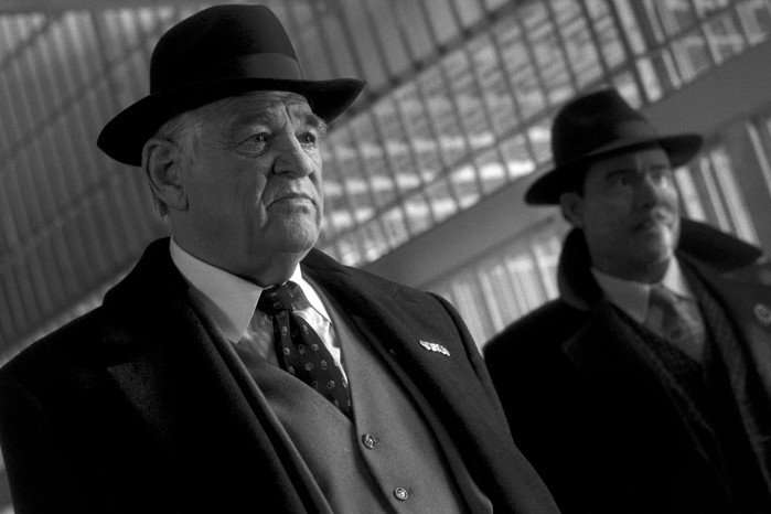 Brendan Gleeson plays Silvermane in Spider-Noir; in this scene, taken from the black-and-white version of the show, he wears a fedora and a tailored suit. He stands, looking very unimpressed, next to a similarly addresses associate.