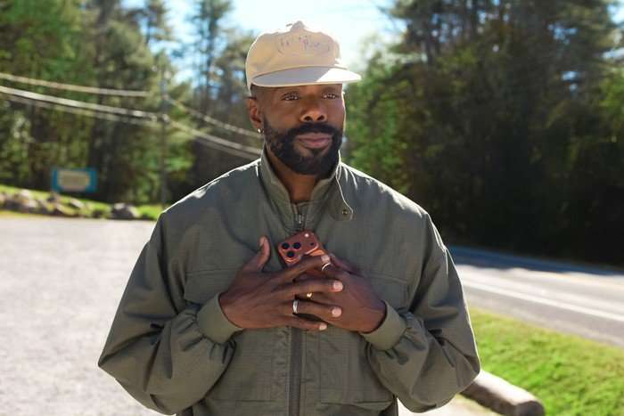 THE FOUR SEASONS, SEASON 2 A bearded man stands outdoors by the roadside on a sunny day, holding a smartphone against his chest with both hands. He is wearing a light-coloured baseball cap, a green zip-up jacket and several rings. Trees and power lines are visible in the softly blurred background, and his expression appears thoughtful and emotional.