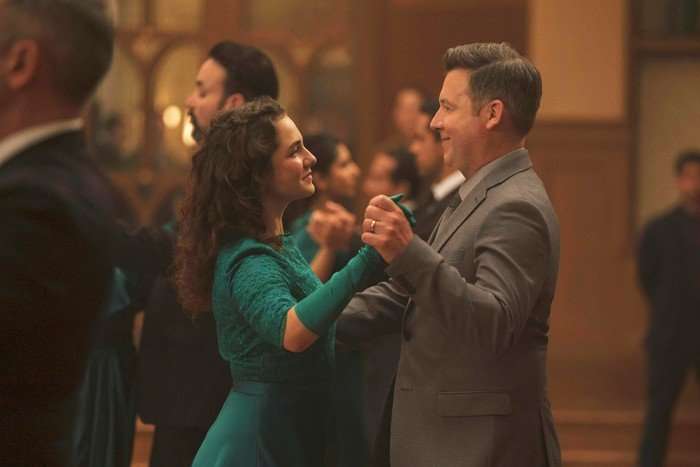 A man and woman are slow dancing together in a warmly lit ballroom or reception hall. The woman has long curly dark hair and is wearing a teal dress with matching gloves, while the man is dressed in a light gray suit and tie. They are smiling softly at each other as they dance, surrounded by other couples blurred in the background, creating an intimate and elegant atmosphere.