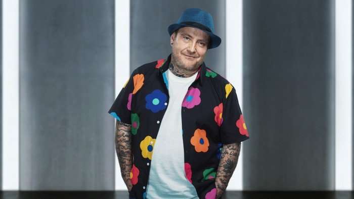 Billy stood wearing a white t-shirt and a black floral shirt with a blue hat, smiling with his hands in his pockets.