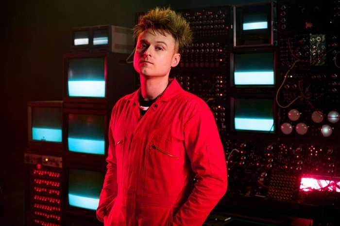 eurovision-sam-battle (1) Sam Battle stood next to a coloured background, wearing a red boiler suit.