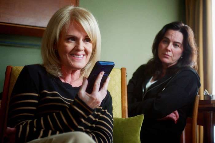 Two women are seated indoors in a cozy, softly lit room. In the foreground, a blonde woman wearing a black sweater with thin horizontal stripes is smiling as she holds a phone or small recording device close to her mouth, as if speaking into it or listening to a message. She appears engaged and slightly amused. In the background, another woman with dark shoulder-length hair sits with her arms crossed, wearing a dark hoodie. She looks toward the blonde woman with a more serious or skeptical expression. The setting includes upholstered chairs, a cushion, and a window with curtains, giving the scene a relaxed, domestic feel.