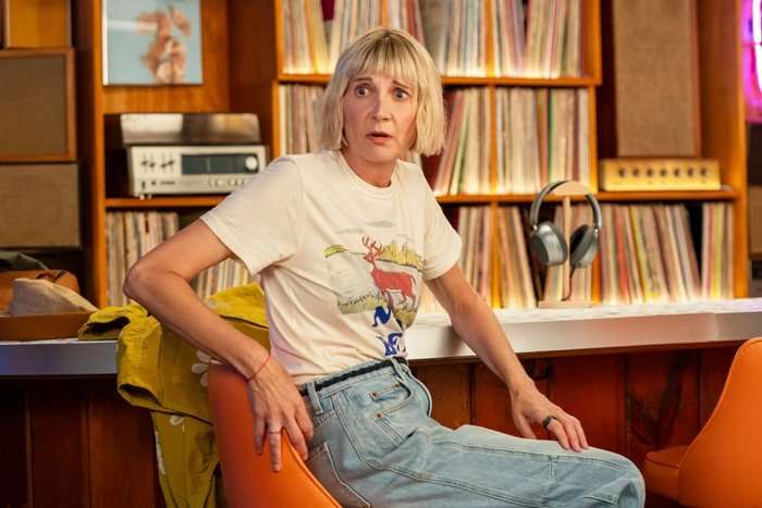 THE FOUR SEASONS, SEASON 2 A blonde woman with a short bob haircut sits in an orange chair in front of shelves filled with vinyl records. Wearing a white graphic T-shirt and light blue jeans, she looks off-camera with a startled expression. The warmly lit room features retro audio equipment, speakers and headphones, creating a cosy music-listening space.