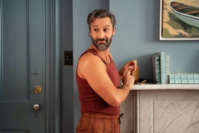 THE FOUR SEASONS, SEASON 2 A bearded man with greying hair stands beside a marble fireplace in a stylish room with blue-grey walls. Wearing a fitted rust-coloured tank top and matching trousers, he glances over his shoulder with an amused expression while holding a small bottle. A framed painting and neatly arranged books sit on the mantelpiece behind him.