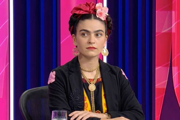 AI-generated depiction of Frida Kahlo seated on the Question Time set, wearing a floral headpiece, gold jewellery and a black jacket against a pink and blue studio backdrop.