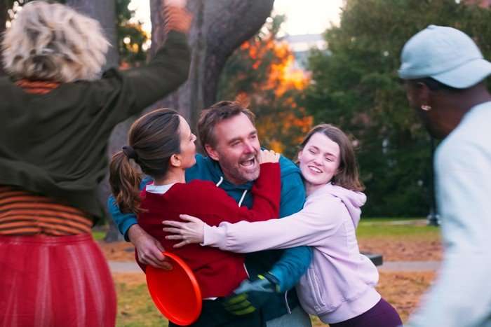 THE FOUR SEASONS A group of friends share a joyful group hug outdoors in a park-like setting at sunset. A man in a blue hoodie stands at the centre, smiling and laughing as two women embrace him from either side. Other figures are partially visible in the foreground, adding to the lively, celebratory atmosphere. Trees and warm evening light fill the background.
