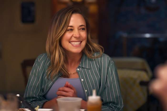 THE FOUR SEASONS, SEASON 2 A smiling woman with shoulder-length blonde hair sits at a dining table in a warmly lit room. She is wearing a blue top layered under a green striped shirt and has her hands clasped together in front of her. Plates, glasses and condiment bottles are visible on the table, while the softly blurred background creates a cosy, intimate atmosphere.