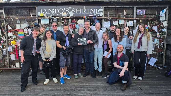 A crowd of filmmakers in front of Ianto's Shrine, with actor Gareth David-Lloyd and Carol-Anne Hillman in the centre holding a clapperboard.