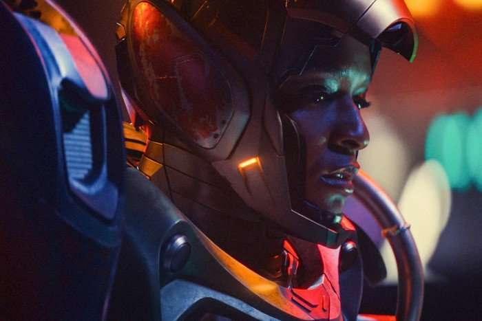 Riri Williams in her Ironheart suit with the faceplate lifted up to reveal her expression, which is one of concern. Multicoloured city lights reflect off the armour.