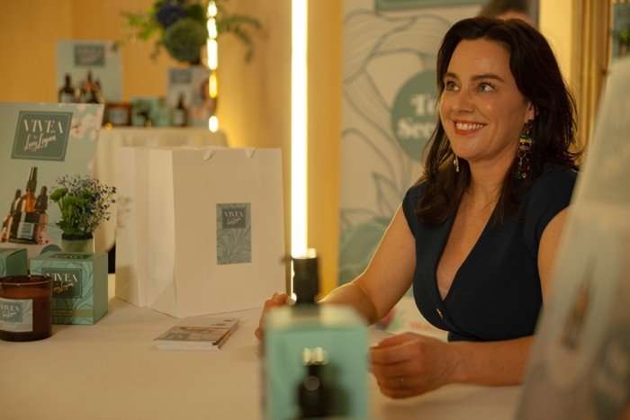 A smiling woman with dark shoulder-length hair sits at a table in a warmly lit indoor setting, likely a market stall or boutique display. She is wearing a navy sleeveless top and colorful dangling earrings. In front of her are neatly arranged skincare or beauty products in teal packaging, along with a white shopping bag featuring a matching label. Behind her, more product displays and signage are visible, suggesting a branded setup. The atmosphere feels inviting and polished, with soft golden lighting and decorative elements like plants and product stands.