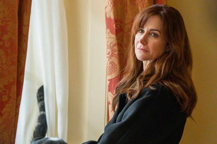 Katherine Kelly in Channel 4 drama Number 10, sitting beside a softly lit window with red patterned curtains as she turns back with a guarded, uneasy expression.