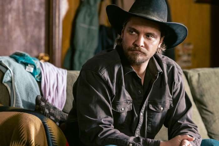 Luke Grimes stars in Marshals: A Yellowstone Story &ndash; he is sat on a couch in a living room, wearing a jacket and a cowboy hat, looking over sternly at someone
