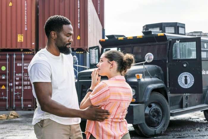 A man gently holds a young woman by the arm as she looks up at him with a worried expression, her hand raised to her mouth. They stand in an industrial area with shipping containers and a large armored truck behind them, suggesting a tense or urgent moment.