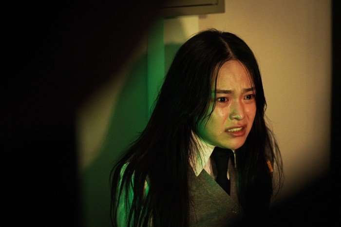 A distressed schoolgirl in a grey uniform looks fearfully off-camera in a dimly lit scene from If Wishes Could Kill, with green light casting across her tear-streaked face.