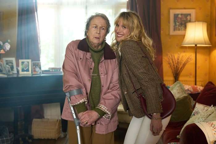 An older woman and a younger woman stand close together in a cozy, warmly lit living room. The older woman, wearing a pink jacket over a green sweater, looks slightly uneasy or puzzled. She is holding a crutch for support, suggesting she may have an injury or mobility issue. Beside her, a smiling blonde woman leans in cheerfully, dressed in a patterned jacket and white pants, with a red shoulder bag. Her upbeat expression contrasts with the older woman&rsquo;s more uncertain look. The background shows a comfortable home setting: a piano with framed photos on top, a glowing floor lamp, curtains filtering daylight, and colorful cushions on a sofa, giving the scene a homely, lived-in feel.