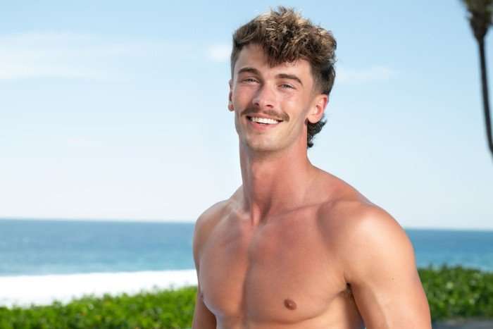 A topless man smiling ahead while stood in front of a beach.