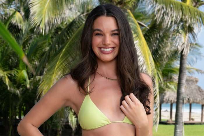 A woman smiling ahead as she stands in front of palm trees, wearing a light green bikini.