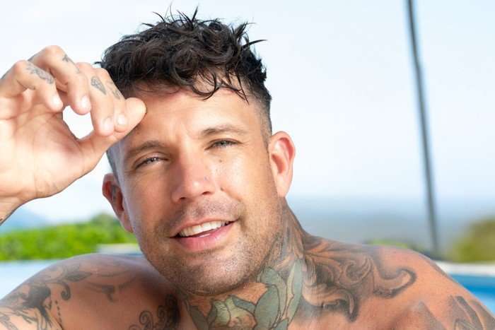 A topless man sat down in front of a swimming pool, smiling ahead with his hand resting on his forehead.