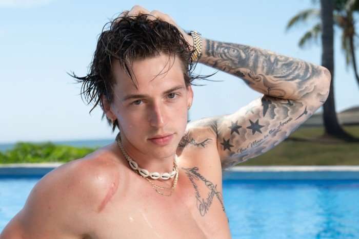 A topless man sat leaning back in front of a swimming pool as his runs his hands through his hair.