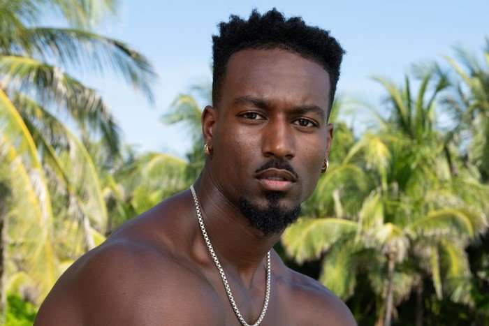 A topless man looking ahead as he sits down in front of palm trees.
