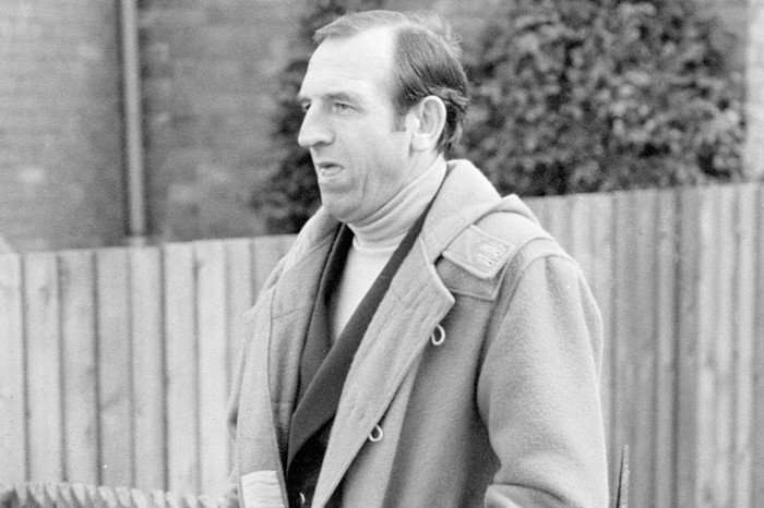 reginald-perrin Black-and-white photo of Leonard Rossiter in The Fall and Rise of Reginald Perrin in a thick coat standing outdoors in front of a wooden fence.