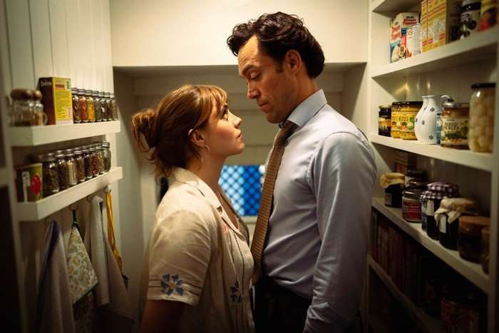 A man and woman stand closely facing each other in a narrow pantry lined with shelves of jars, spices and kitchen items. The woman, wearing a light embroidered blouse with her hair tied back, looks up at the man, who is dressed in a shirt and tie. Warm lighting and their intense expressions create a tense, intimate atmosphere.