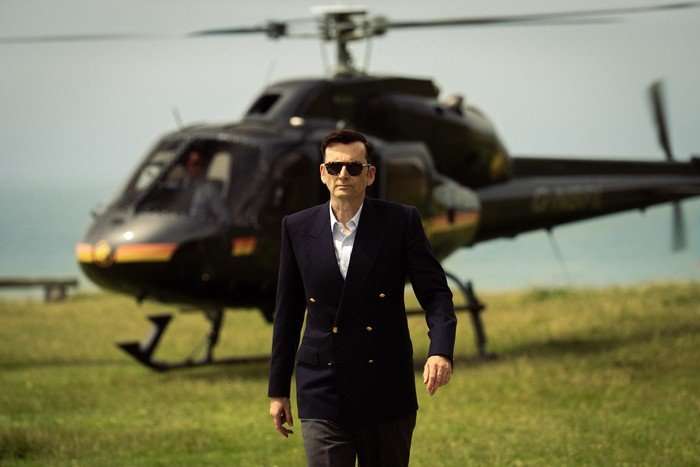 David Tennant as Lord Tony Baddingham in Rivals season 2. He is seen here wearing formal clothing and walking away from a helicopter landed on a grass lawn