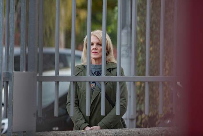 A blonde woman stands behind a tall metal gate with vertical bars, which partially obscure her face and body. She appears to be outdoors, possibly on a street or near a residential area&mdash;there&rsquo;s a parked car and some greenery visible in the background. She&rsquo;s wearing a green coat over a grey sweater and has a crossbody bag strap across her chest. Her posture is upright, hands together in front of her, and her expression looks serious or concerned as she gazes forward through the bars. The framing gives a sense of separation or confinement due to the gate in the foreground.